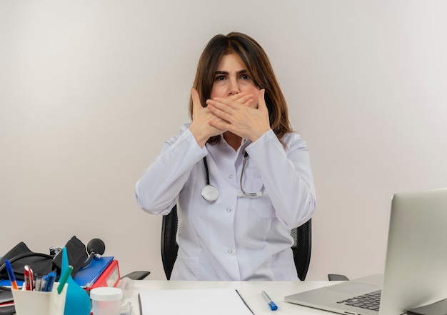 Medico femminile di mezza età interessato che indossa la veste medica con lo stetoscopio che si siede alla scrivania lavora sul computer portatile con strumenti medici ha coperto la bocca con le mani sul muro bianco con lo spazio della copia