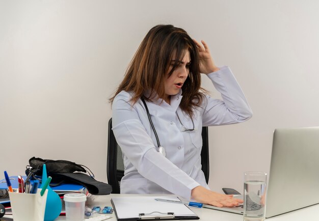 Обеспокоенная женщина-врач средних лет в медицинском халате со стетоскопом, сидя за столом, работает на ноутбуке с медицинскими инструментами, использовала ноутбук, положив руку на голову на белой стене с копией пространства