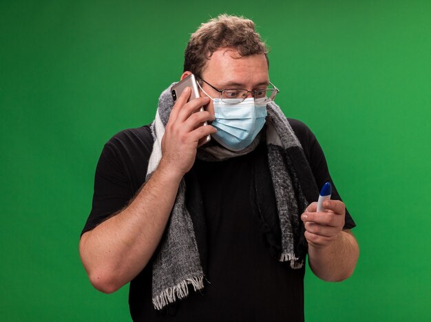 Concerned middle-aged ill male wearing medical mask and scarf speaks on phone and looking at thermometer in his hand 