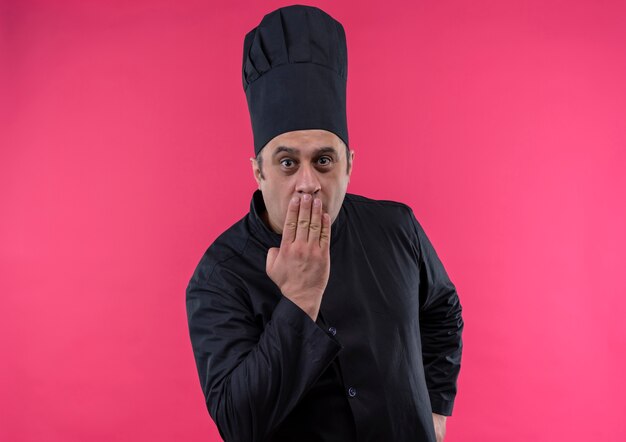 Concerned middle-aged male cook in chef uniform covered mouth with hand on isolated pink wall