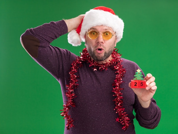 Concerned middle-aged man wearing santa hat and tinsel garland around neck with glasses holding christmas tree toy with date keeping on head looking at camera isolated on green background