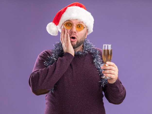 보라색 벽에 고립 된 얼굴에 손을 유지 샴페인 잔을 들고 안경 목에 산타 모자와 반짝이 화환을 입고 우려 중년 남자