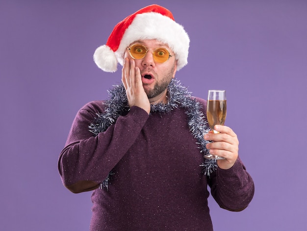 Concerned middle-aged man wearing santa hat and tinsel garland around neck with glasses holding glass of champagne keeping hand on face  isolated on purple wall