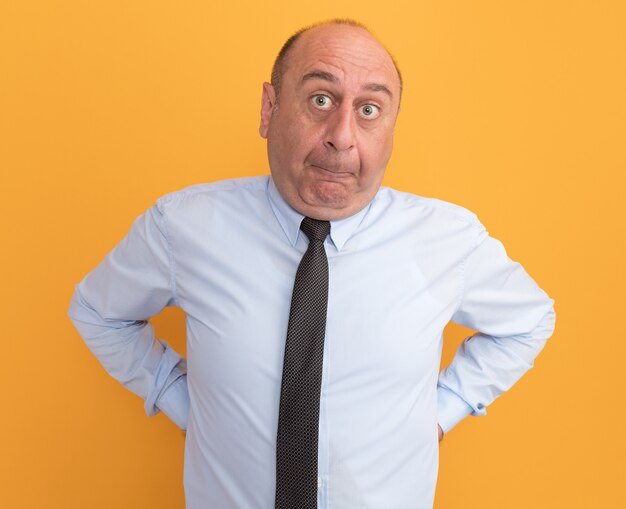 Concerned middle-aged man wearing white t-shirt with tie putting hands on waist isolated on orange wall