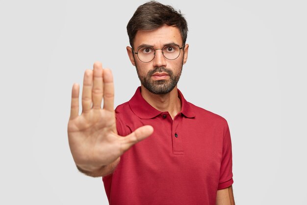 Concerned serious bearded man in round eyewear pulls palm towards camera, stops or warns you