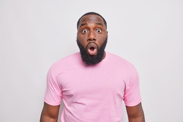 Concerned shocked bearded man stares bugged eyes at front opens mouth in amazement reacts on surprising news dressed in casual pink t shirt isolated over white wall