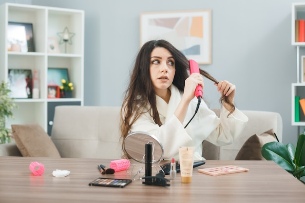 Обеспокоенная выпрямить волосы с помощью плоского железа молодая девушка сидит за столом с инструментами для макияжа в гостиной