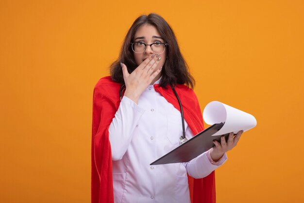 concerned superhero girl in red cape wearing doctor uniform and stethoscope with glasses holding clipboard looking at front putting hand on mouth 