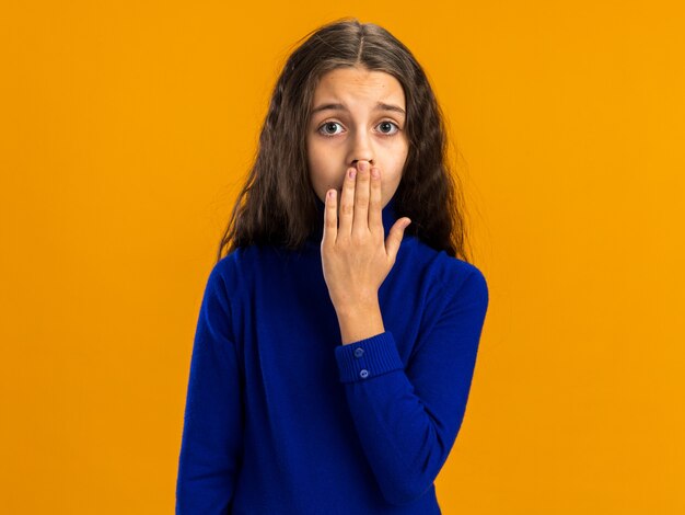 Concerned teenage girl keeping hand on mouth  isolated on orange wall with copy space