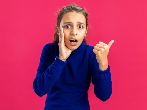 Concerned teenage girl  keeping hand near mouth whispering pointing at side isolated on pink wall