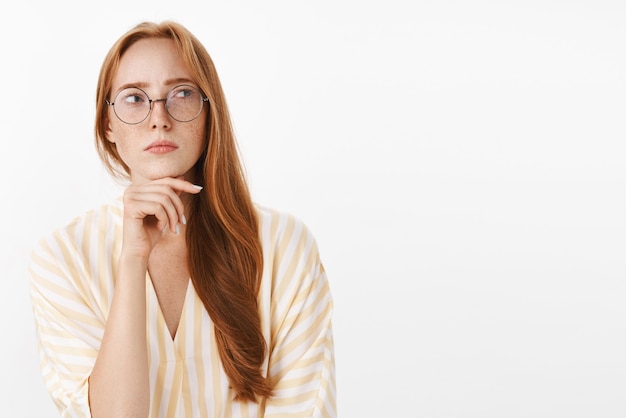 Concerned thoughtful and concentrated creative female writer with red hair and freckles in glasses and trendy yellow blouse standing in hmm pose looking right doubtful and focused touching chin