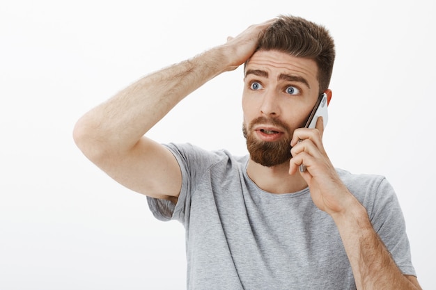 Concerned and troubled anxious boyfriend receiving bad news during phone call holding arm on forehead looking left concerned and clueless standing confused and perplexed over gray wall