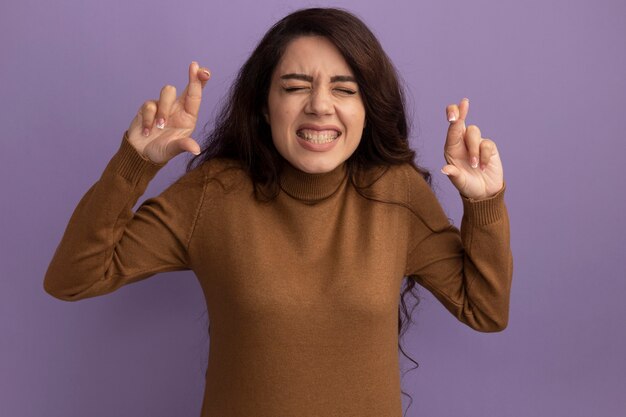 Concerned with closed eyes young beautiful girl wearing brown turtleneck sweater crossing fingers isolated on purple wall