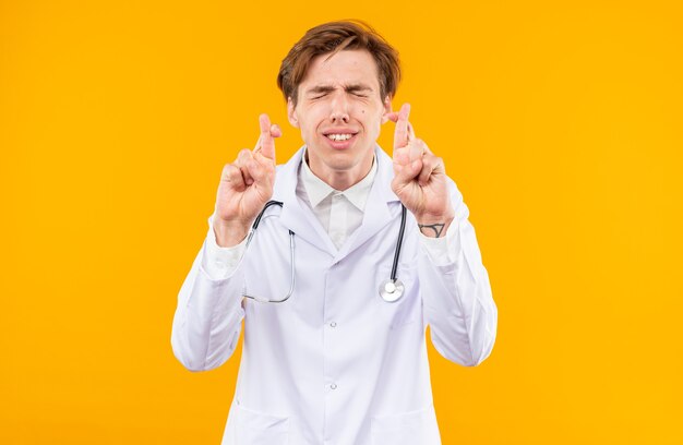 Concerned with closed eyes young male doctor wearing medical robe with stethoscope crossing fingers isolated on orange wall