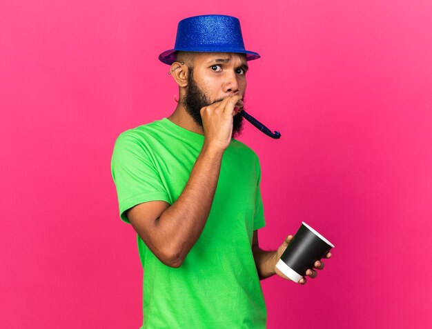 분홍색 벽에 격리된 커피 한 잔을 들고 파티 모자를 쓰고 있는 걱정스러운 젊은 아프리카계 미국인 남자