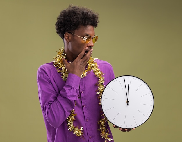 Concerned young afro-american man wearing glasses with tinsel garland around neck holding and looking at clock keeping hand on mouth isolated on olive green background