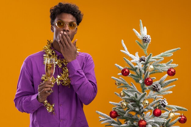 Concerned young afro-american man wearing glasses with tinsel garland around neck standing near decorated christmas tree holding glass of champagne keeping hand