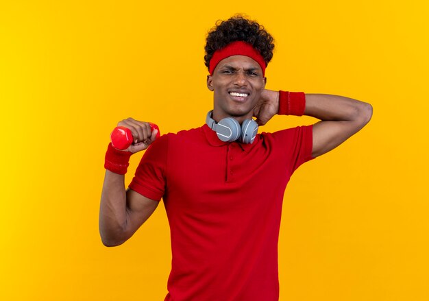 Concerned young afro-american sporty man wearing headband and wristband with headphones on neck holding dumbbell and putting hand behind head isolated on yellow wall