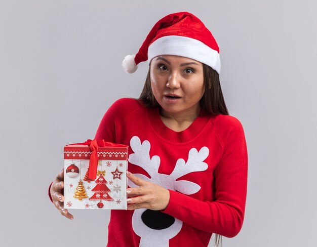 Обеспокоенная молодая азиатская девушка в новогодней шапке со свитером, держащая подарочную коробку на белом фоне