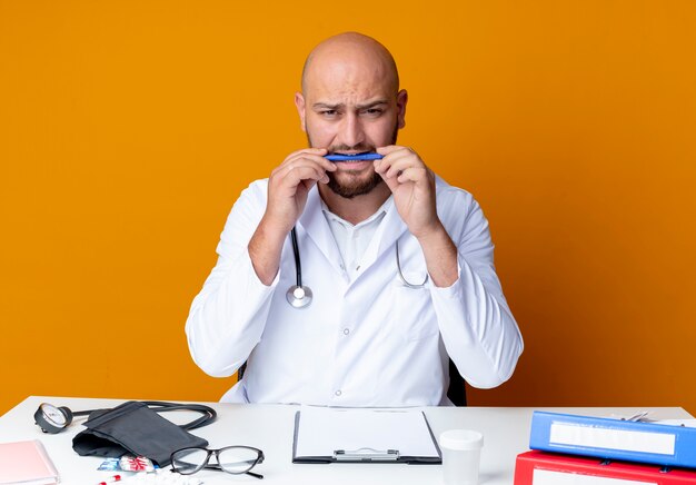 Concerned young bald male doctor wearing medical robe and stethoscope sitting at work desk with medical tools bites pen isolated on orange background