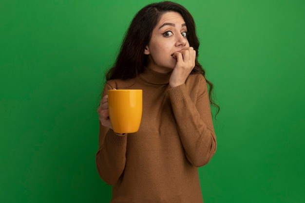 Обеспокоенная молодая красивая девушка, держащая чашку чая, кусает ногти, изолированные на зеленой стене