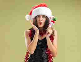 無料写真 オリーブグリーンの背景で隔離のクリスマスのおもちゃを保持している首に花輪とクリスマスの帽子をかぶって心配している若い美しい少女