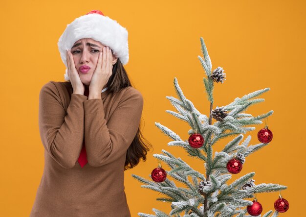 Concerned young beautiful girl wearing christmas hat with tie standing nearby christmas tree putting hands on cheeks isolated on orange background