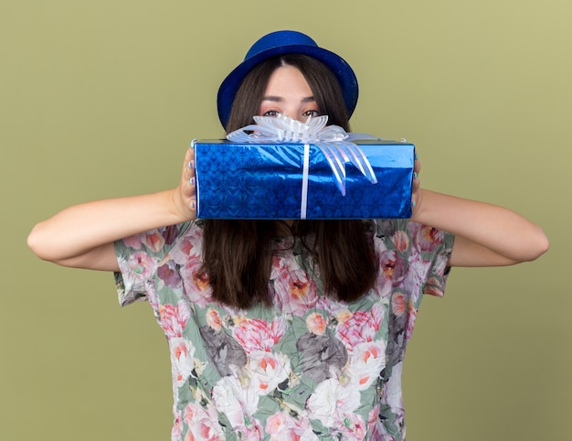 La giovane bella ragazza interessata che indossa il cappello da festa ha coperto il viso con una confezione regalo isolata su una parete verde oliva