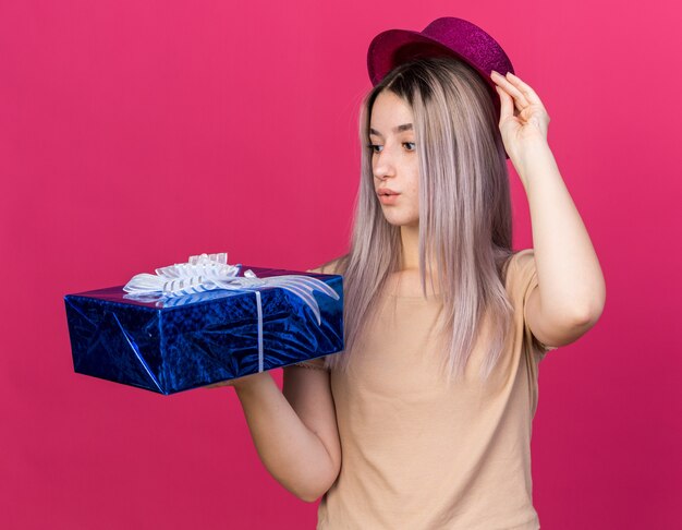 Concerned young beautiful girl wearing party hat holding and looking at gift box 