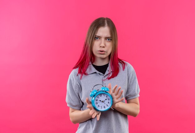 孤立したピンクの壁に目覚まし時計を保持している灰色のTシャツを着て心配している若い美しい女性