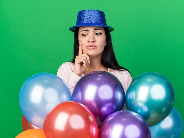 Concerned young beautiful woman wearing party hat standing behind balloons putting finger on cheek isolated on green wall
