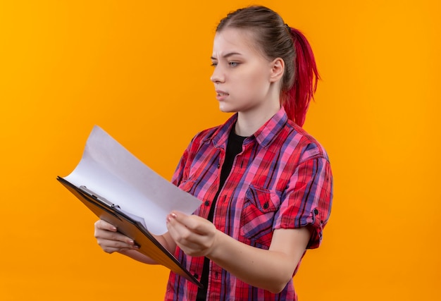 Concerned young beautiful woman wearing red shirt flipping through clipboard on isolated yellow wall