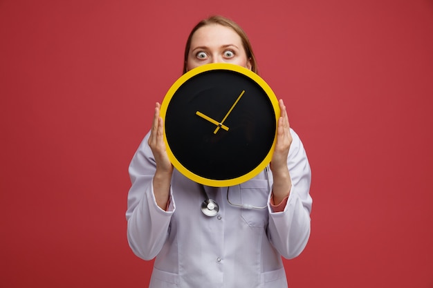 Concerned young blonde female doctor wearing medical robe and stethoscope around neck holding clock from behind it 