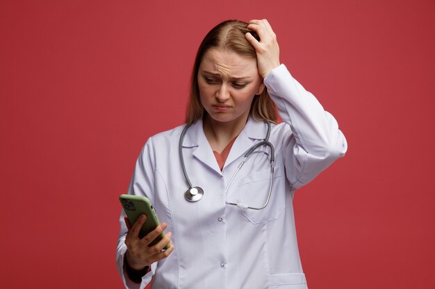 Concerned young blonde female doctor wearing medical robe and stethoscope around neck keeping hand on head holding and looking at mobile phone 