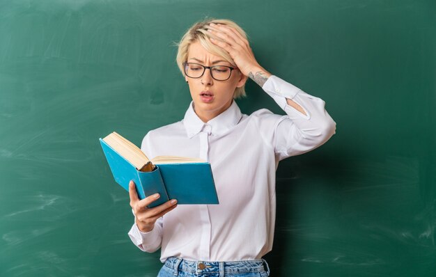 Concerned young blonde female teacher wearing glasses in classroom standing in front of chalkboard keeping hand on head holding and reading book