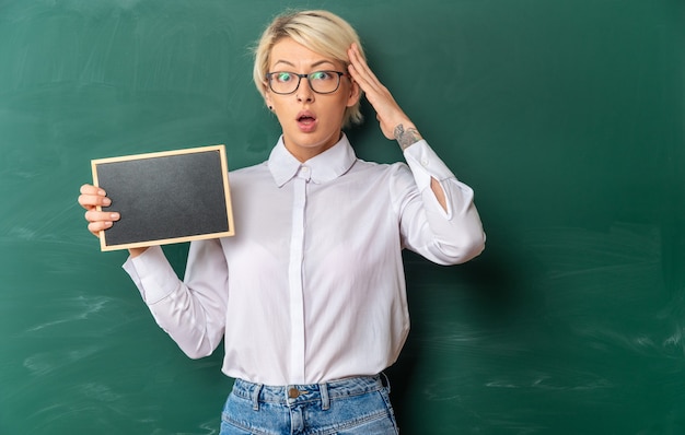 concerned young blonde female teacher wearing glasses in classroom standing in front of chalkboard showing mini blackboard keeping hand on head looking at front with copy space