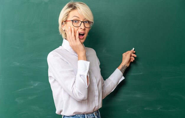 心配している若い金髪の女教師が教室で眼鏡をかけて黒板の前に立って黒板を指さし、チョークで正面を向いて手を顔に向けている