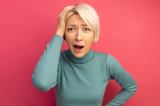 Concerned young blonde girl keeping hand on waist and on head looking  isolated on pink wall