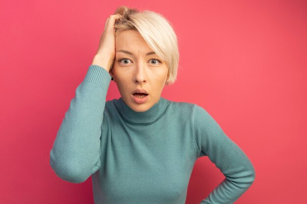 Concerned young blonde girl keeping hand on waist and on head looking  isolated on pink wall