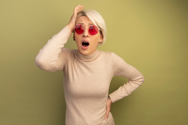 Free photo concerned young blonde girl wearing sunglasses putting hand on head keeping another one on waist looking at side