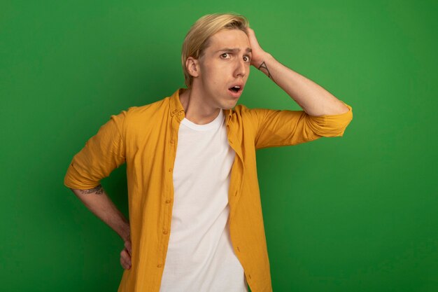 Обеспокоенный молодой блондин в желтой футболке, положив руку на голову, изолированную на зеленом