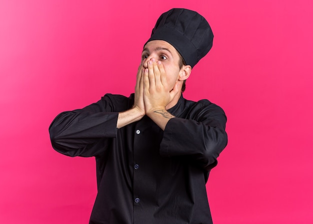 Concerned young blonde male cook in chef uniform and cap covering mouth with hands looking at side isolated on pink wall