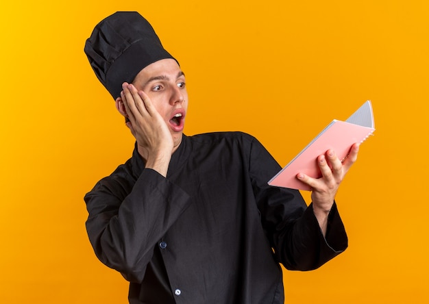 Concerned young blonde male cook in chef uniform and cap keeping hand on face reading note pad isolated on orange wall