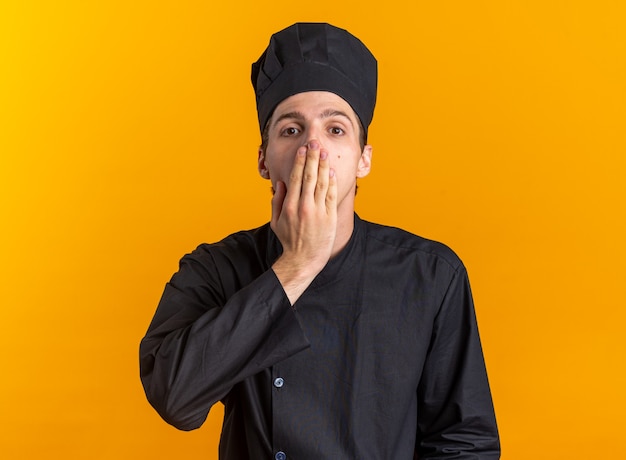 Обеспокоенный молодой блондин мужчина-повар в униформе шеф-повара и кепке смотрит в камеру, прикрывая рот рукой, изолированной на оранжевой стене