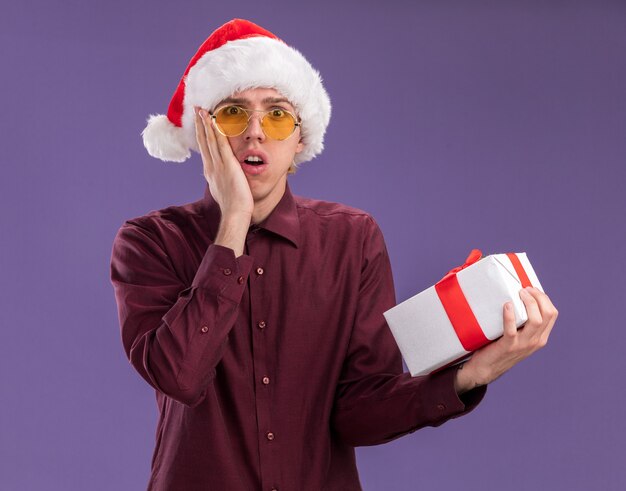 Concerned young blonde man wearing santa hat and glasses holding gift package looking at camera keeping hand on face isolated on purple background