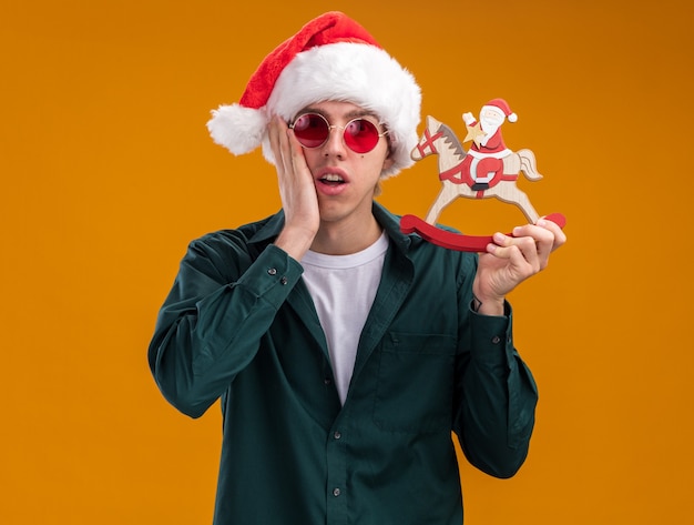 Concerned young blonde man wearing santa hat and glasses holding santa on rocking horse figurine looking at camera keeping hand on face isolated on orange background