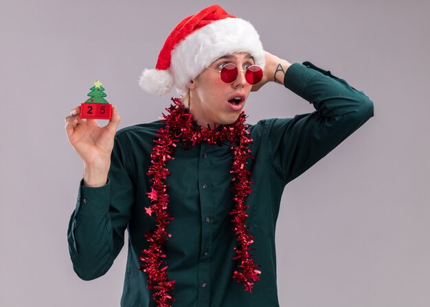 흰색 배경에 고립 된 측면을보고 머리에 손을 유지하는 날짜와 함께 크리스마스 트리 장난감을 들고 목 주위에 반짝이 갈 랜드와 산타 모자와 안경을 착용 우려 젊은 금발의 남자