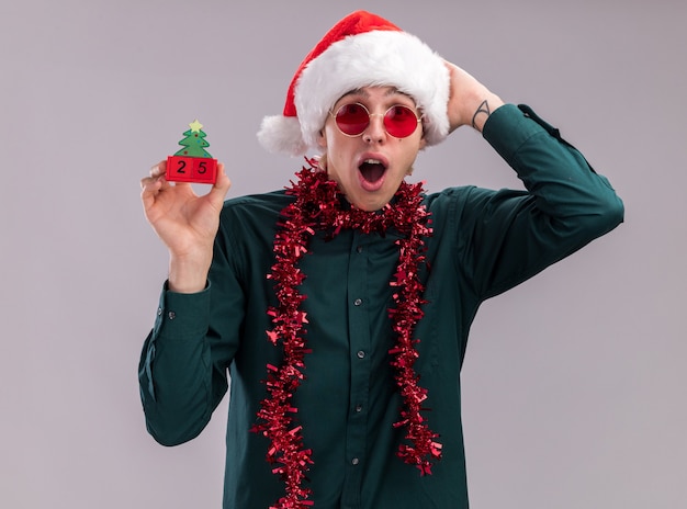 サンタの帽子とメガネを身に着けている心配している若いブロンドの男が首の周りに見掛け倒しの花輪を持ってクリスマスツリーのおもちゃを保持している