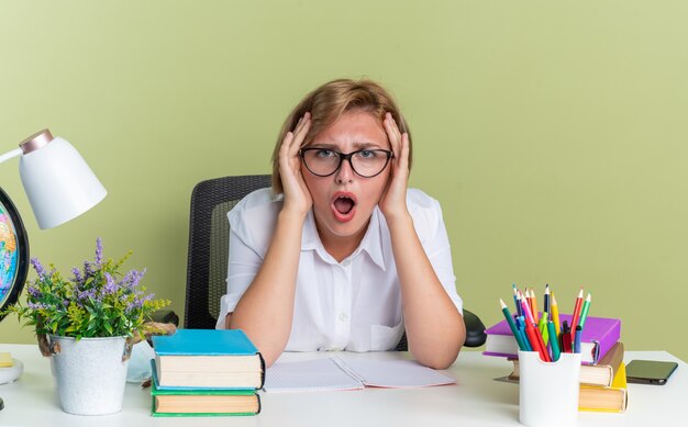 Concerned young blonde student girl wearing glasses sitting at desk with school tools keeping hands on head looking at camera isolated on olive green wall