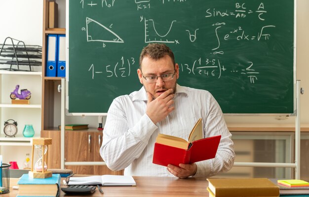 Concerned young blonde teacher wearing glasses sitting at desk with school tools in classroom reading book keeping hand on chin